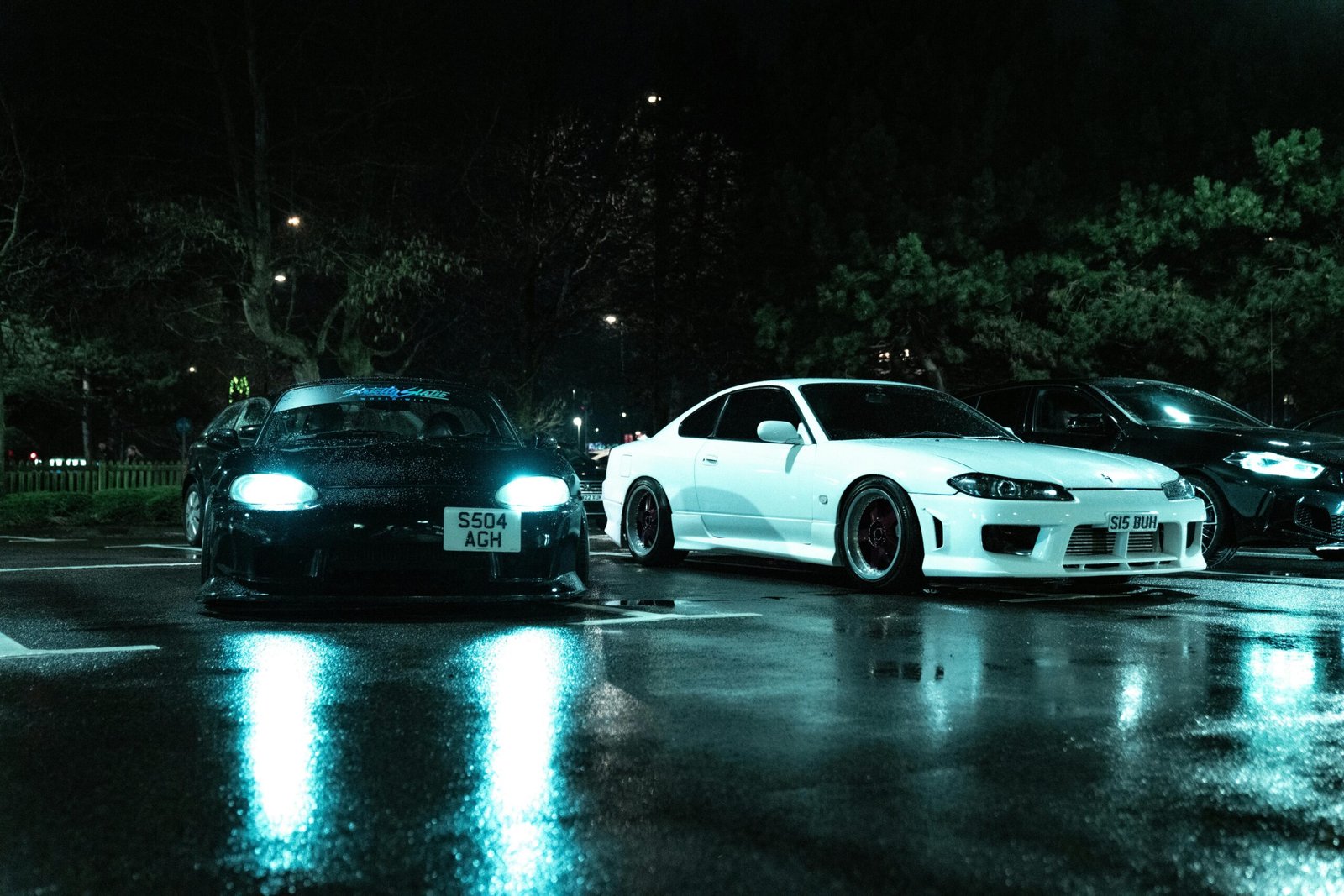 Two Japanese sports cars parked in a wet urban lot at night, with a moody atmosphere.